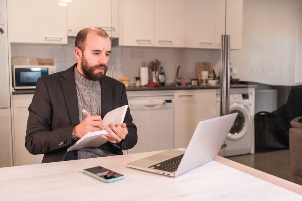 Mann in einem Anzug, der in einem Notizbuch schreibt, während er auf einen Laptop schaut.